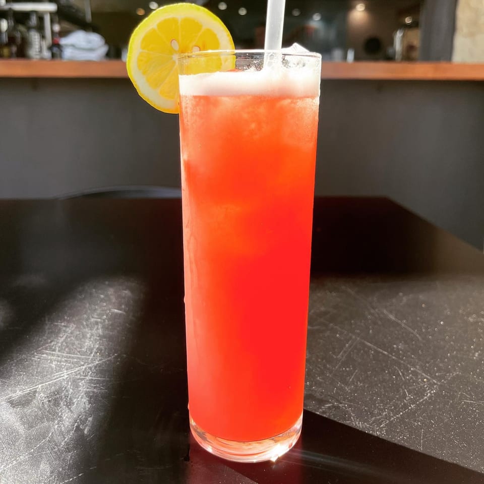A tall glass of red drink with a lemon slice and straw on a dark table at Harold Dean Smoked Goods.