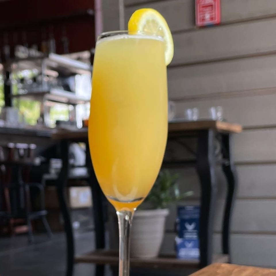 A mimosa cocktail with a lemon slice garnish, set against a blurred restaurant background.