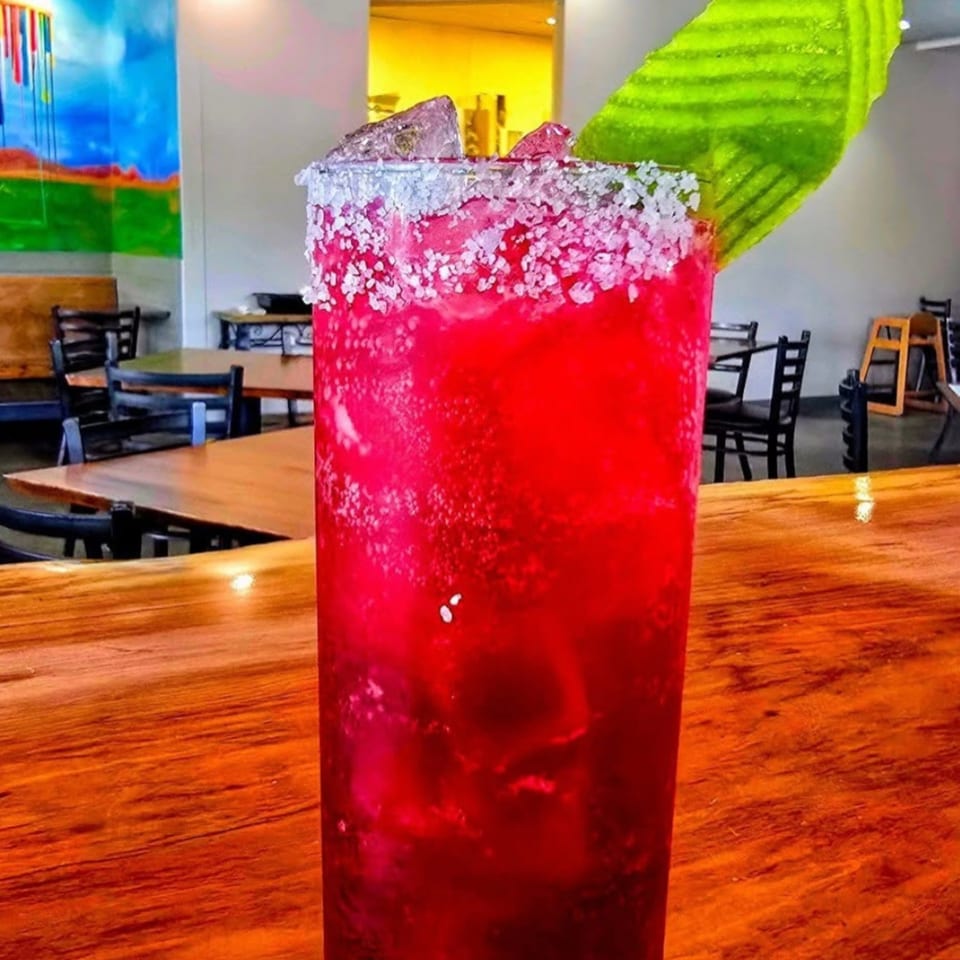 A vibrant red cocktail with a salted rim and green garnish sits on a wooden bar top.
