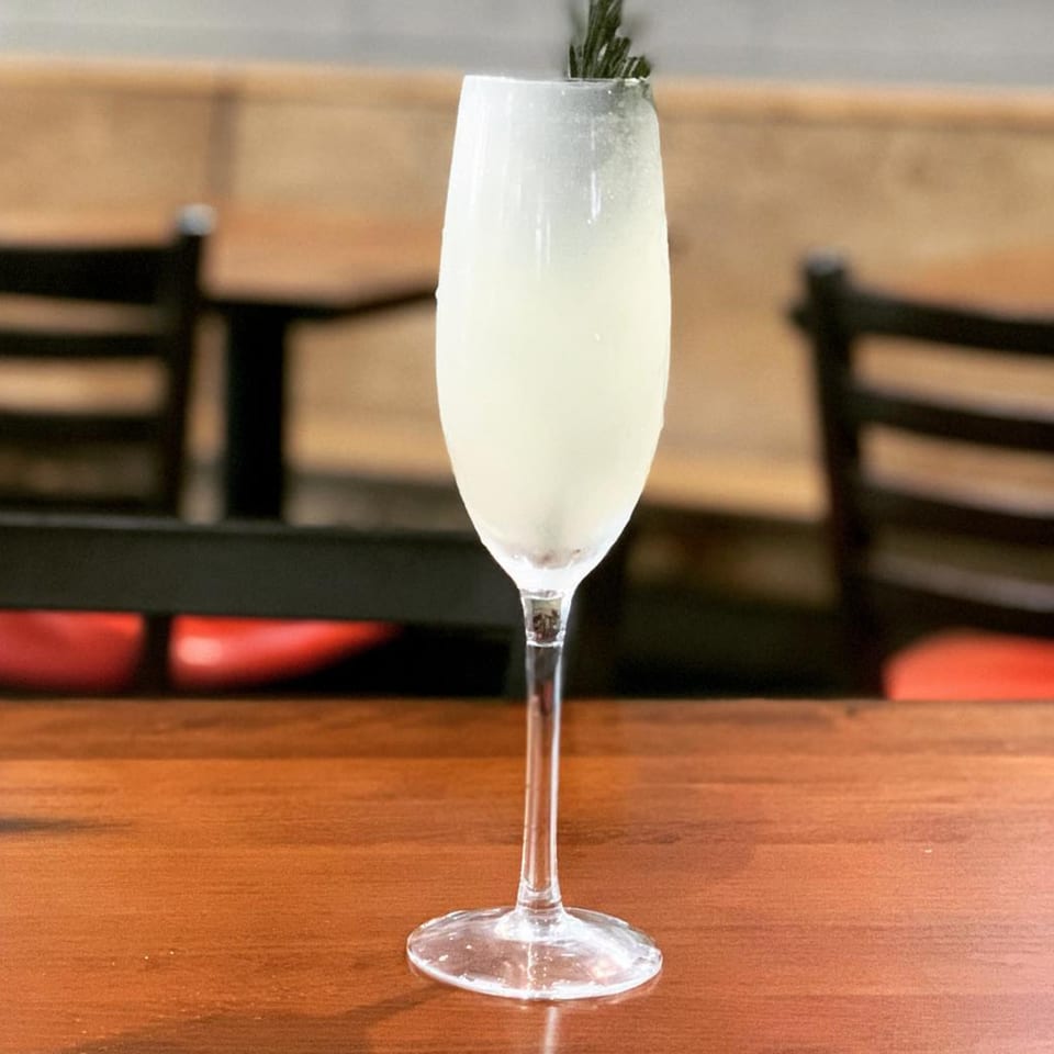 A tall, clear glass of light-colored cocktail with a dark garnish sits on a wooden table.